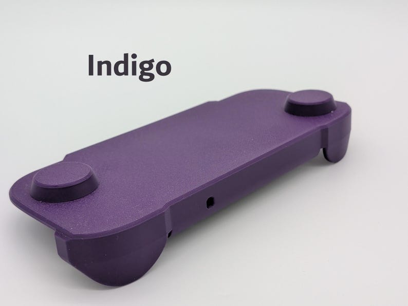 May include: A purple handheld gaming controller grip with a textured matte finish. The device features two circular thumbstick areas on the top surface and a small port on the side. The word Indigo is printed in dark gray text above the controller.