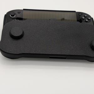 Pu&ograve; includere: Una console di gioco portatile nera con una cover nera strutturata. La cover ha due sporgenze circolari e una forma rettangolare. La console ha uno schermo e pulsanti su entrambi i lati.