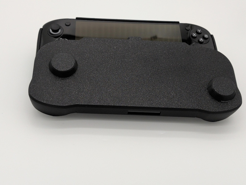 Pu&ograve; includere: Una console di gioco portatile nera con una cover nera strutturata. La cover ha due sporgenze circolari e una forma rettangolare. La console ha uno schermo e pulsanti su entrambi i lati.