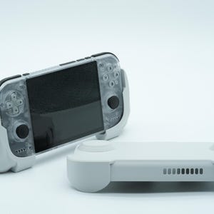 Pode incluir: Uma consola de jogos portátil com caixa transparente e botões pretos, apresentando um ecrã central e controlos integrados. Ao lado encontra-se uma pega de proteção ou caixa de comando cinzento-claro com lado texturado e ranhuras.