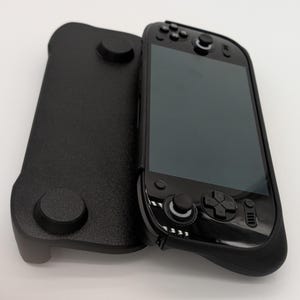 Pu&ograve; includere: Un dispositivo di gioco portatile nero con una custodia protettiva nera abbinata. Il dispositivo presenta uno schermo, pulsanti e joystick. La custodia ha due sporgenze circolari e una superficie strutturata.