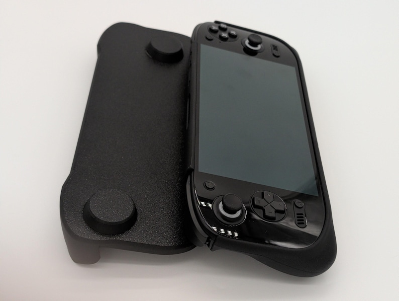 Pu&ograve; includere: Un dispositivo di gioco portatile nero con una custodia protettiva nera abbinata. Il dispositivo presenta uno schermo, pulsanti e joystick. La custodia ha due sporgenze circolari e una superficie strutturata.