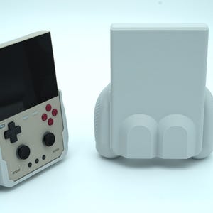 Puede incluir: Una consola de juegos portátil con placa frontal de color beige claro, pantalla negra, pad direccional negro, dos joysticks negros y cuatro botones de acción rojos, dentro de una funda de agarre gris claro texturizada, junto a un soporte a juego.