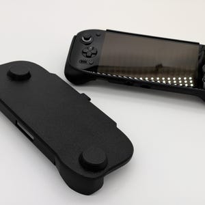 Pu&ograve; includere: Due console di gioco portatili nere e una custodia protettiva nera sono mostrate su sfondo bianco. Le console hanno uno schermo, pulsanti e joystick. La custodia ha due elementi circolari in rilievo.