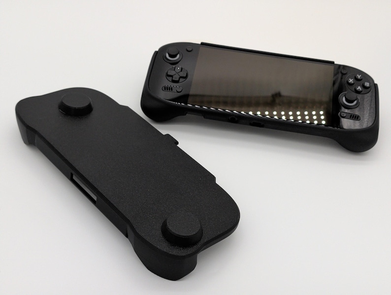 Pu&ograve; includere: Due console di gioco portatili nere e una custodia protettiva nera sono mostrate su sfondo bianco. Le console hanno uno schermo, pulsanti e joystick. La custodia ha due elementi circolari in rilievo.