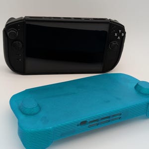 May include: A black handheld gaming console with a screen, directional pad, analogue sticks, and X, Y, A, B buttons, positioned behind a separate, translucent teal protective cover that features cutouts for the device's ports and controls.