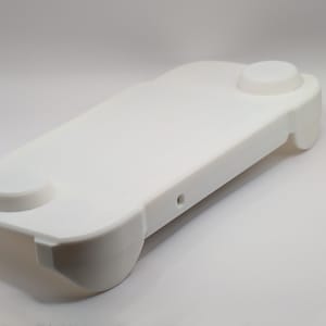 May include: A solid white, rectangular plastic controller grip accessory with rounded edges. The top surface features two circular raised buttons on opposite ends and a small, centered circular port opening on the side edge of the frame.