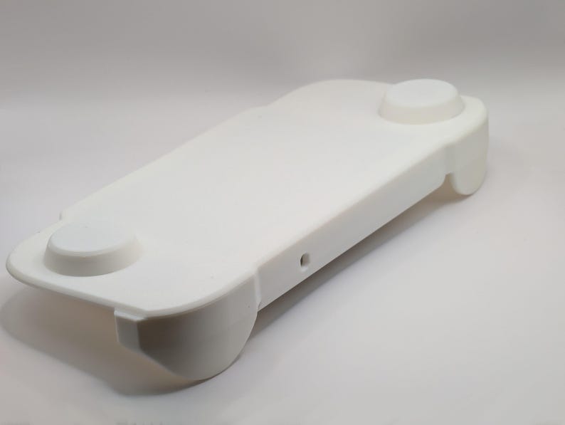 May include: A solid white, rectangular plastic controller grip accessory with rounded edges. The top surface features two circular raised buttons on opposite ends and a small, centered circular port opening on the side edge of the frame.