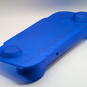 May include: A bright blue 3D-printed handheld gaming controller grip featuring a rectangular base with rounded edges, two circular raised thumbstick cutouts on the top surface, and ergonomic side handles for a comfortable grip during gameplay.