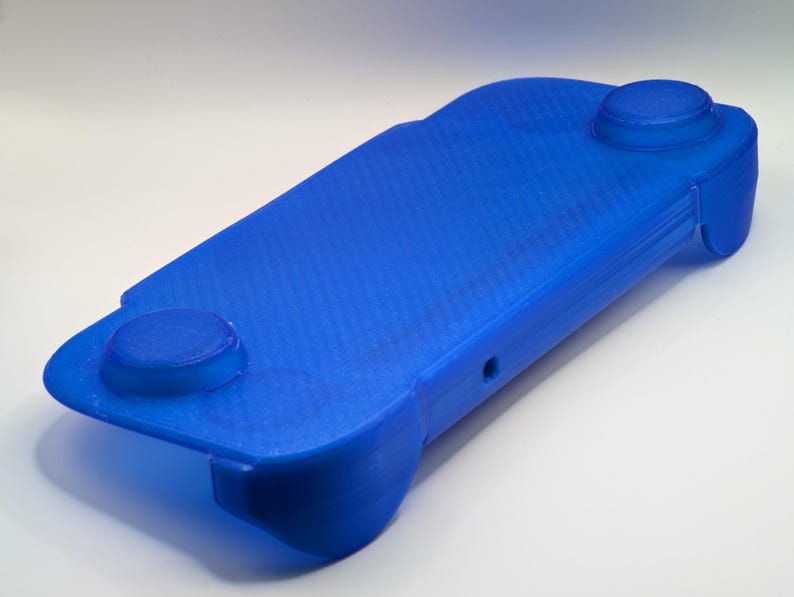May include: A bright blue 3D-printed handheld gaming controller grip featuring a rectangular base with rounded edges, two circular raised thumbstick cutouts on the top surface, and ergonomic side handles for a comfortable grip during gameplay.