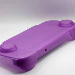 Puede incluir: Un dispositivo de juego portátil de color púrpura con dos botones circulares y una superficie texturizada. El dispositivo tiene bordes redondeados y un diseño elegante. El color es un púrpura vibrante y sólido.