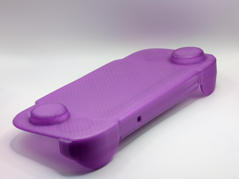 May include: A purple 3D-printed handheld gaming console grip featuring a rectangular central body with two integrated circular thumbstick covers on the top surface and contoured ergonomic handles on each side for a comfortable grip.