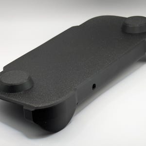 May include: A black, rectangular, textured plastic controller grip accessory featuring two circular raised thumbstick covers on the top surface and contoured side handles designed for ergonomic support during handheld gaming sessions.