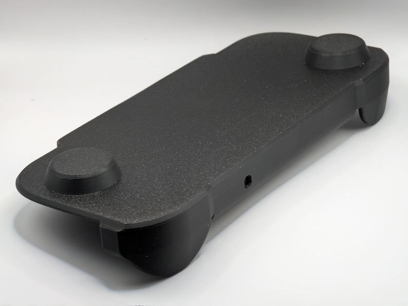 May include: A black, rectangular, textured plastic controller grip accessory featuring two circular raised thumbstick covers on the top surface and contoured side handles designed for ergonomic support during handheld gaming sessions.