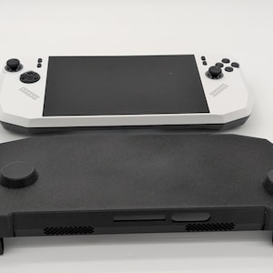 以下が含まれることがあります： 黒い画面、アナログスティック、ボタンを備えた白い携帯ゲーム機と、その手前に配置された黒い保護カバー。カバーにはアナログスティック用の円形の切り抜きと、下端に沿った通気口のグリルが施されています。