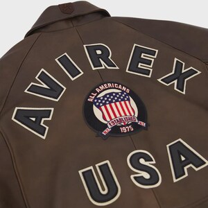 May include: Brown leather jacket with the word "AVIREX" across the back in white lettering with black outlines. A circular patch with the American flag and the words "ALL AMERICANS ESTABLISHED 1975" is also visible. The word "USA" is below the patch.