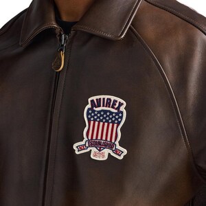 May include: Brown leather jacket with a zip-up front and a classic collar. The jacket features a patch on the chest with the text "AVIREX NY ESTABLISHED 1975" in a patriotic design with red and white stripes and blue stars.