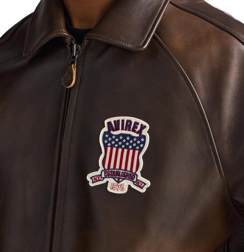May include: Brown leather jacket with a zip-up front and a classic collar. The jacket features a patch on the chest with the text "AVIREX NY ESTABLISHED 1975" in a patriotic design with red and white stripes and blue stars.