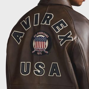 May include: Brown leather jacket with the word "AVIREX" across the back in white lettering. A circular patch with the American flag and the words "ALL AMERICANS ESTABLISHED 1975" is centered below the lettering. The word "USA" is below the patch.