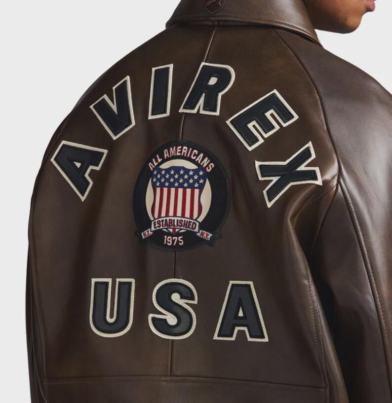 May include: Brown leather jacket with the word "AVIREX" across the back in white lettering. A circular patch with the American flag and the words "ALL AMERICANS ESTABLISHED 1975" is centered below the lettering. The word "USA" is below the patch.