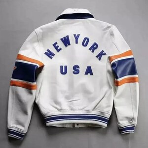 Puede incluir: Chaqueta de cuero blanca con detalles en azul y naranja. La parte trasera presenta el texto "NEW YORK USA" en azul. Las mangas tienen rayas azules y naranjas. La chaqueta tiene un dobladillo y pu&ntilde;os a rayas azules y blancas.