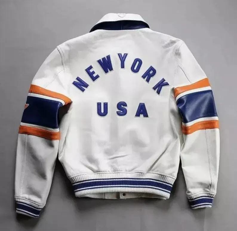 Puede incluir: Chaqueta de cuero blanca con detalles en azul y naranja. La parte trasera presenta el texto "NEW YORK USA" en azul. Las mangas tienen rayas azules y naranjas. La chaqueta tiene un dobladillo y pu&ntilde;os a rayas azules y blancas.