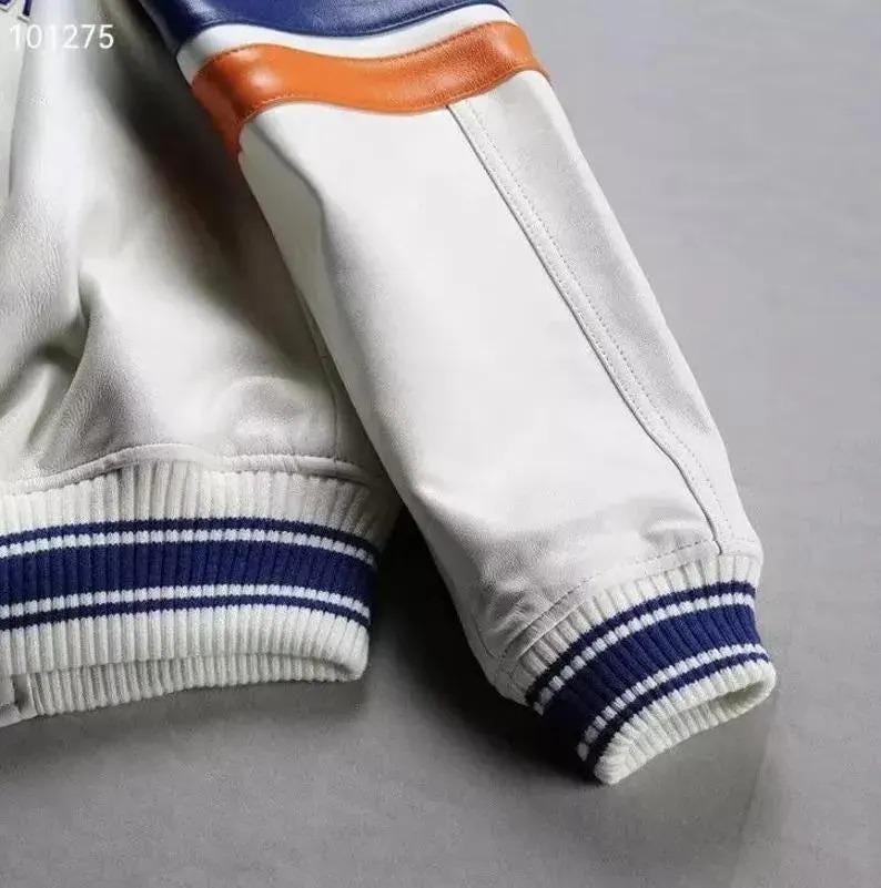 Puede incluir: Primer plano de una manga de chaqueta de cuero blanca con detalles azules y naranjas. El pu&ntilde;o es acanalado con rayas azules y blancas. La chaqueta tiene un estilo deportivo y atl&eacute;tico.
