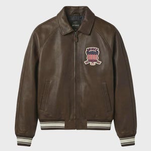 May include: Brown leather jacket with a collar and a zipper closure. The jacket features a patch with a red, white, and blue design. The cuffs and hem have white and cream stripes. The brand name "AVIREX" is visible on the jacket.