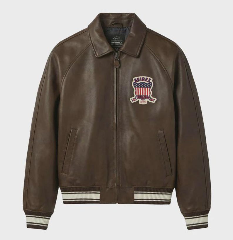 May include: Brown leather jacket with a collar and a zipper closure. The jacket features a patch with a red, white, and blue design. The cuffs and hem have white and cream stripes. The brand name "AVIREX" is visible on the jacket.