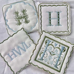Puede incluir: Cuatro posavasos blancos bordados con bordes festoneados. Cada posavasos presenta un diseño diferente: una letra "H" adornada, las iniciales "HWL" y dos diseños "H" más. El bordado es en tonos verdes y azules.