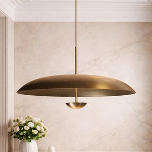 Può includere: Una moderna lampada a sospensione in ottone con un grande paralume poco profondo a forma di disco. La lampada è appesa a un'asta sottile fissata a un supporto a soffitto circolare. Il design è minimalista ed elegante, adatto a vari stili di interni.