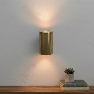 May include: A cylindrical brass wall sconce emits warm light, mounted on a textured gray wall. The sconce is positioned above a wooden surface, with a small potted succulent and stacked books. The lighting fixture adds a touch of elegance to the space.