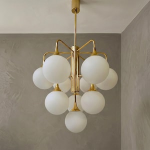 Op de afbeelding: Een moderne kroonluchter met een goudkleurig frame en tien witte, bolvormige glazen kappen. De armatuur hangt aan het plafond en is ontworpen om sfeerverlichting te bieden. Het ontwerp is eigentijds en geschikt voor diverse interieurstijlen.