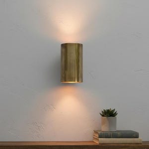 May include: A cylindrical brass wall sconce emits warm light, mounted on a textured white wall. The sconce is positioned above a wooden shelf holding books and a small potted succulent, creating a cozy ambiance.