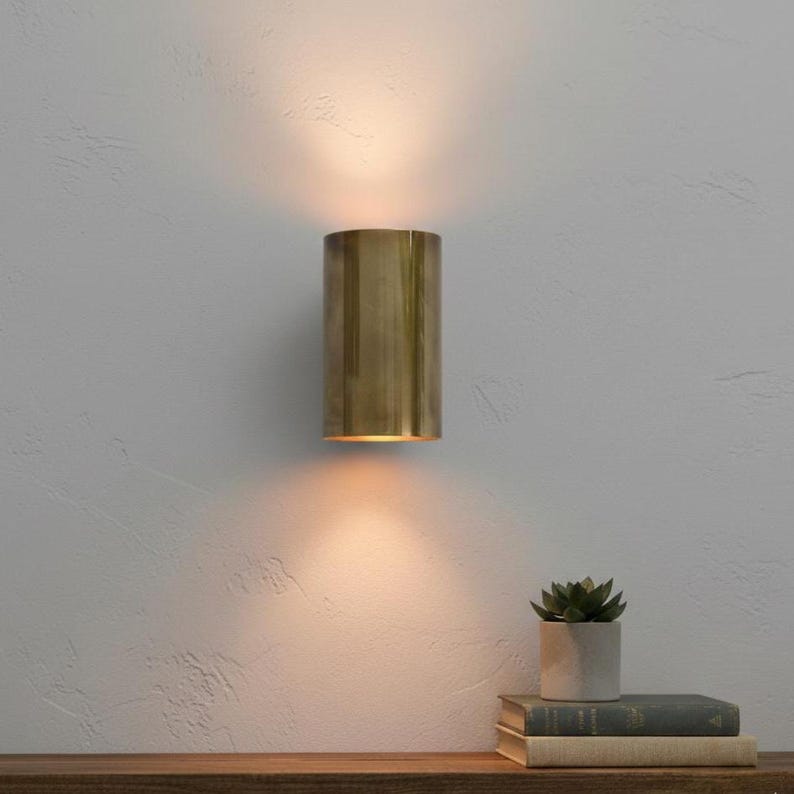 May include: A cylindrical brass wall sconce emits warm light, mounted on a textured white wall. The sconce is positioned above a wooden shelf holding books and a small potted succulent, creating a cozy ambiance.