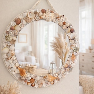 Puede incluir: Espejo redondo decorado con diversas conchas marinas, perlas y un borde de cuerda. El espejo refleja una habitación con tonos neutros y una cesta. El espejo está colgado en una pared blanca.