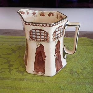 May include: A cream-colored ceramic pitcher featuring a brown-toned illustration of monks in robes walking past arched windows. The rim is decorated with a pattern of hanging bells, and the handle is accented with a dark green trim.
