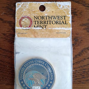 May include: A coin from the Northwest Territorial Mint, packaged in a clear plastic sleeve. The coin features the text "UNITED STATES CYBER COMMAND" around the edge, with an eagle and shield design in the center. The packaging has the text "NORTHWEST TERRITORIAL MINT".