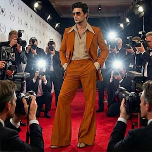 May include: A man in a burnt orange suit with wide-leg pants poses on a red carpet. The suit has a notched lapel and a light tan shirt underneath. Photographers with cameras surround him, with bright flashes illuminating the scene.