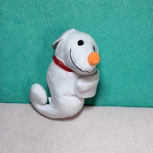 May include: A small white plush toy shaped like a ghost with a rounded orange nose, black embroidered eyes, and a wide black stitched smile. The figure features a red collar around its neck and a curved tail-like base, crafted from soft fabric.