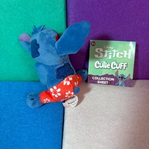 Puede incluir: Un peluche azul del personaje Stitch con pantalones rojos de estampado floral blanco, junto a una hoja de colecci&oacute;n cuadrada verde de Disney Stitch Cutie Cuff con texto. El peluche tiene orejas grandes y una peque&ntilde;a etiqueta blanca.