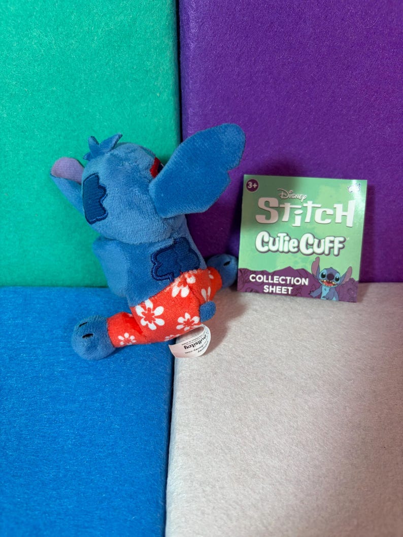 Puede incluir: Un peluche azul del personaje Stitch con pantalones rojos de estampado floral blanco, junto a una hoja de colecci&oacute;n cuadrada verde de Disney Stitch Cutie Cuff con texto. El peluche tiene orejas grandes y una peque&ntilde;a etiqueta blanca.