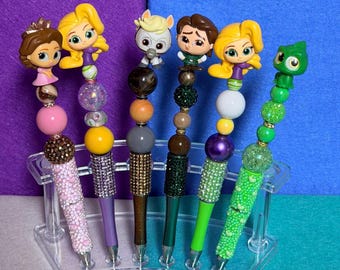 May include: A collection of decorative pens featuring character toppers. The pens have various colors, including pink, purple, brown, green, and yellow. Each pen has a beaded design and a rhinestone-covered barrel. The pens are displayed in a clear plastic stand.
