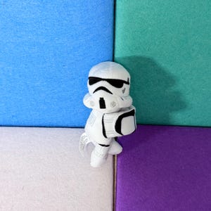 May include: A plush toy shaped like a white stormtrooper helmet with black embroidered details for the eyes, mouth, and armor accents. The soft, stuffed character features a rounded head and a simplified, compact design with visible stitching.