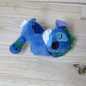 May include: A blue plush toy featuring a character with closed eyes, a dark blue nose, and green accents on the ears and feet. The toy has a white collar, a dark blue flower-shaped patch on the back, and a small white tag attached to the side.