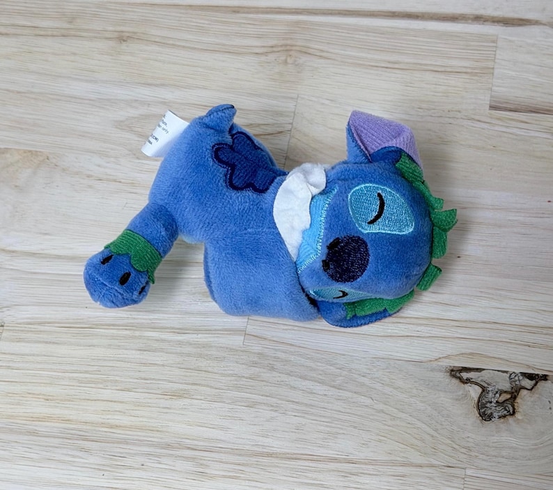 May include: A blue plush toy featuring a character with closed eyes, a dark blue nose, and green accents on the ears and feet. The toy has a white collar, a dark blue flower-shaped patch on the back, and a small white tag attached to the side.