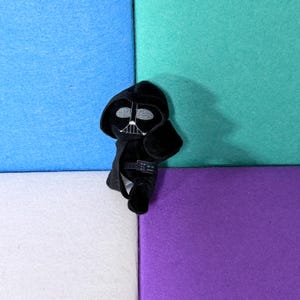 May include: A small, black plush toy character featuring a hooded cape, a mask with silver-stitched eye lenses, and a detailed chest control panel. The figure is constructed from soft fabric and displays a stylized, simplified design of a dark armored figure.