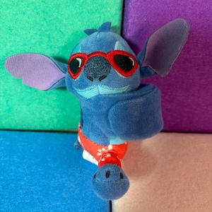Puede incluir: Un peluche azul de un personaje con orejas grandes, que lleva gafas de sol con montura roja y un ba&ntilde;ador rojo con estampado floral blanco. El juguete tiene una textura suave, rasgos faciales bordados y una forma corporal redondeada.