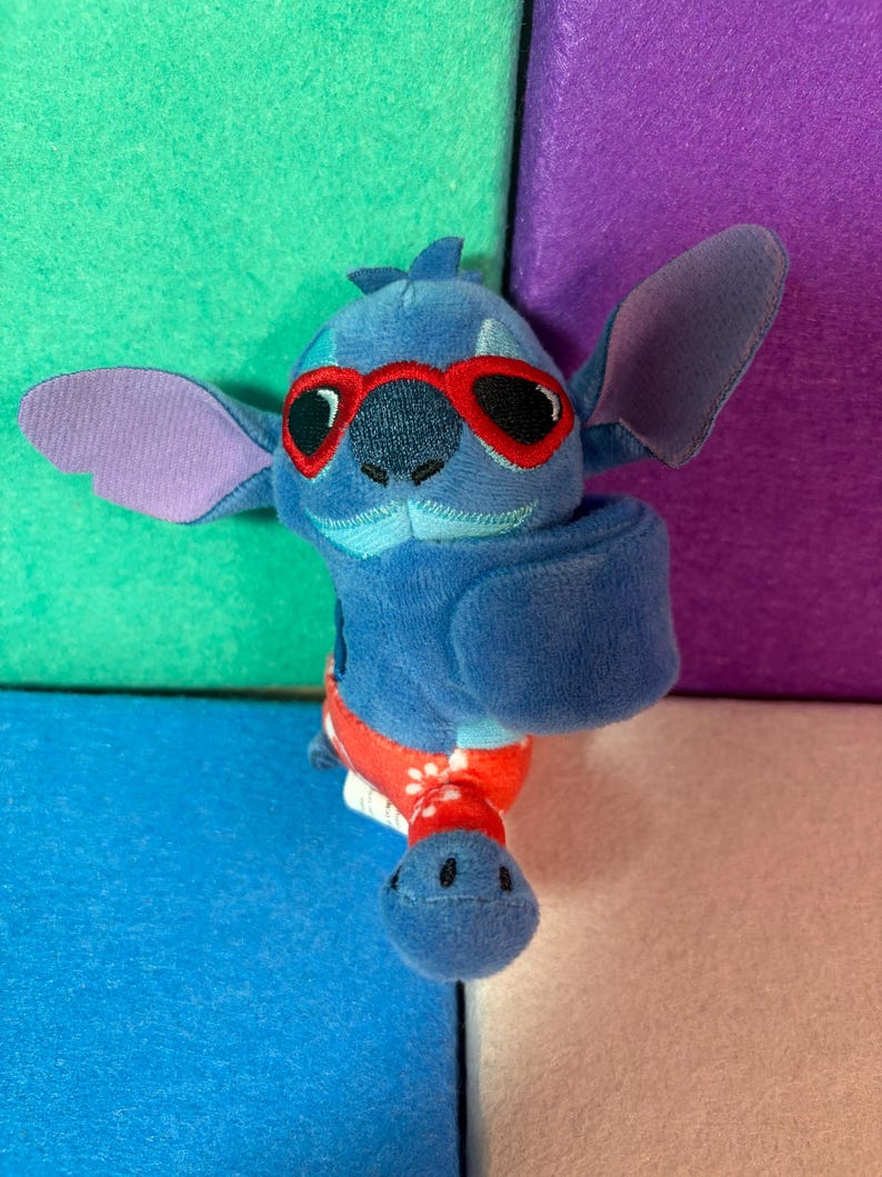 Puede incluir: Un peluche azul de un personaje con orejas grandes, que lleva gafas de sol con montura roja y un ba&ntilde;ador rojo con estampado floral blanco. El juguete tiene una textura suave, rasgos faciales bordados y una forma corporal redondeada.