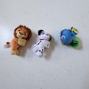 Op de afbeelding: Drie pluche speelgoed: een leeuwenwelp met bruine en bruine vacht, een Dalmatische puppy met zwarte vlekken en een blauwe vis met gele en groene accenten. Elk speelgoed heeft een label. Het speelgoed is ontworpen voor kinderen.
